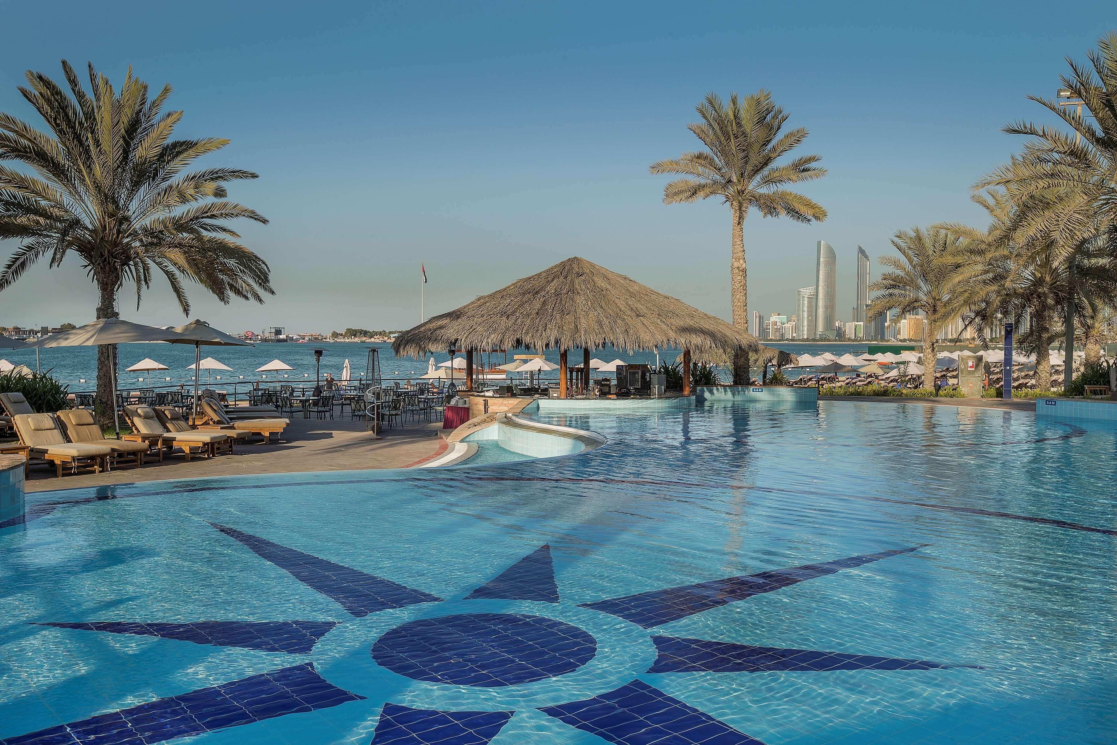 Radisson Blu Hotel & Resort, Abu Dhabi Corniche Exterior photo The photo shows a panoramic view of a luxurious resort pool area. In the foreground, there is a large blue swimming pool with a decorative star pattern on the bottom. Surrounding the pool are several lounge chairs, and there are palm trees providing 