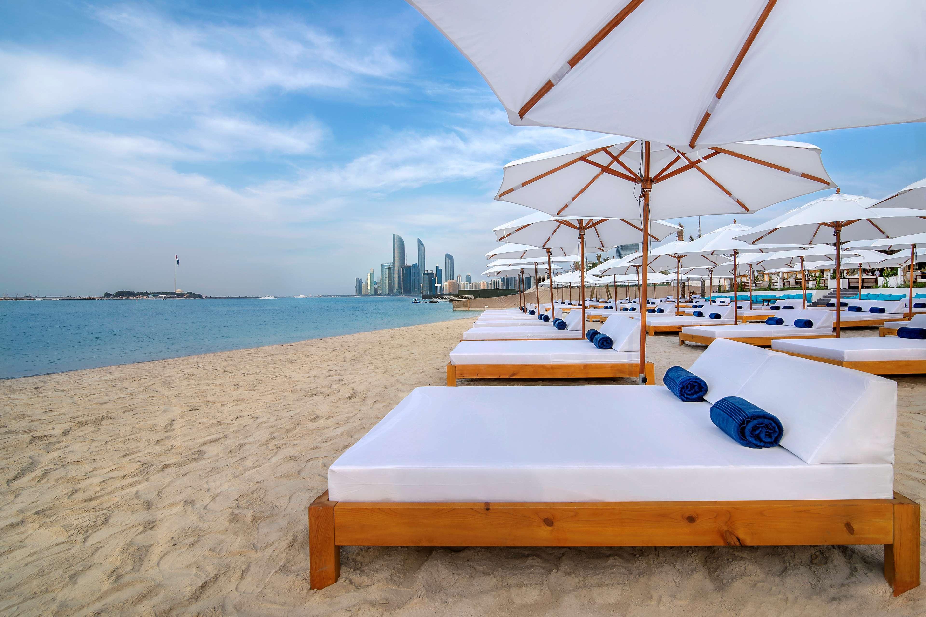 Radisson Blu Hotel & Resort, Abu Dhabi Corniche Exterior photo The photo depicts a serene beach setting with several white lounge beds arranged neatly on the sandy shore. Each bed is paired with a white umbrella providing shade. In the background, a skyline of modern buildings can be seen along the coast, hintin