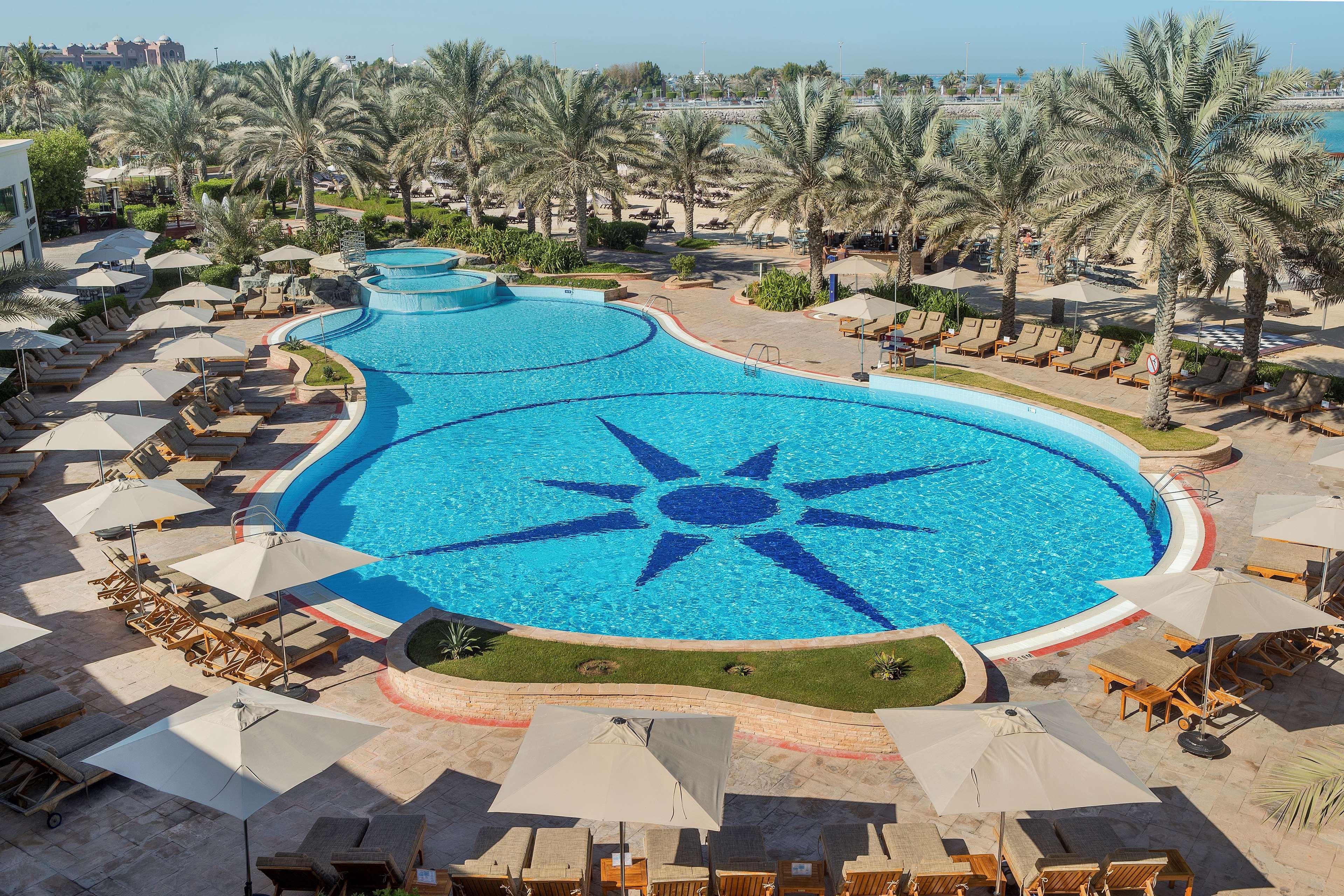 Radisson Blu Hotel & Resort, Abu Dhabi Corniche Exterior photo The image depicts a large, beautifully designed swimming pool shaped like a freeform design, featuring a prominent star pattern at its center. Surrounding the pool are numerous lounge chairs with light-colored cushions and umbrellas, providing shade.