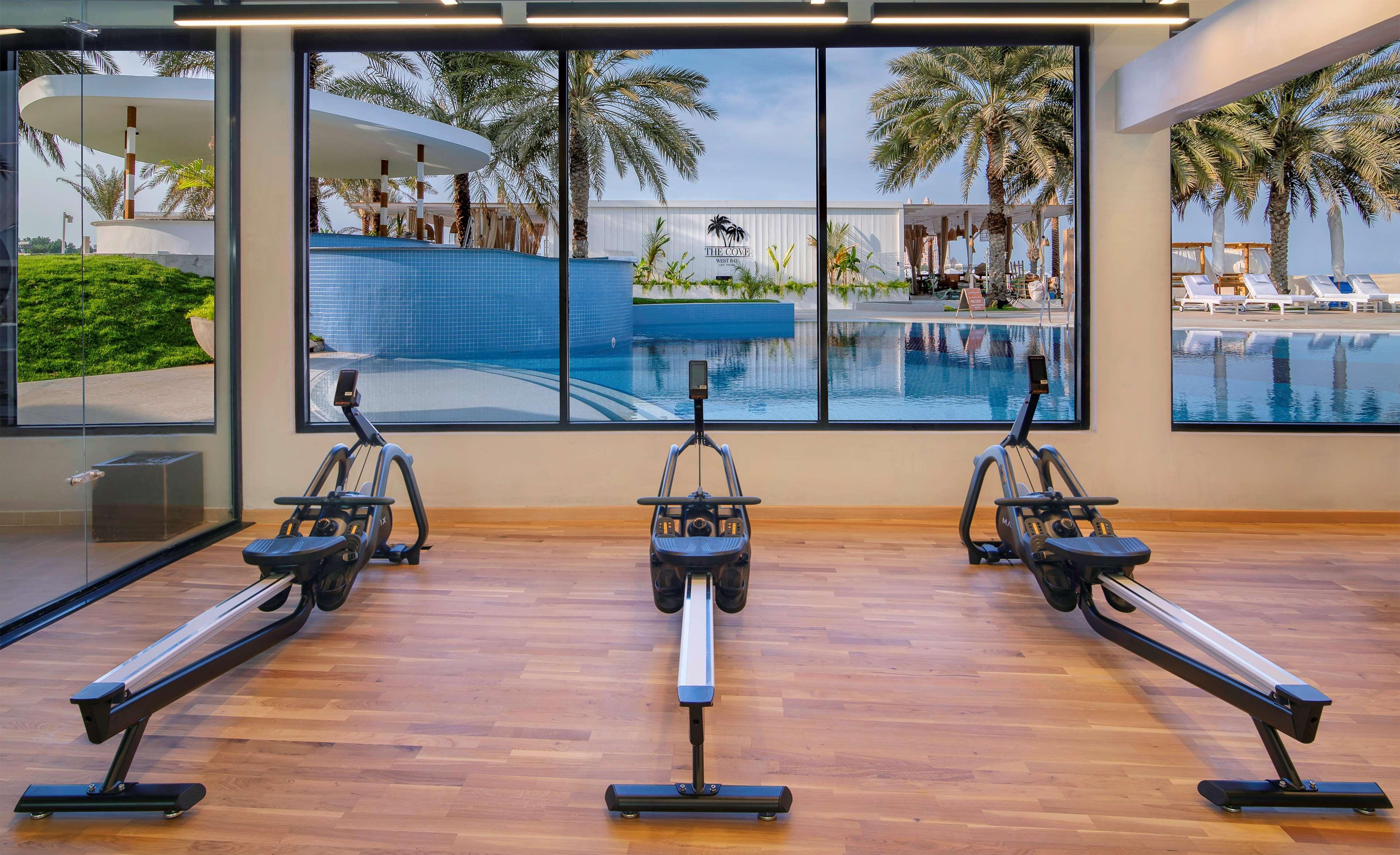 Radisson Blu Hotel & Resort, Abu Dhabi Corniche Exterior photo The photo shows a gym or fitness area featuring three rowing machines lined up on a wooden floor. In the background, there is a large window that displays an outdoor scene with palm trees and a swimming pool, creating a bright and inviting atmosphere