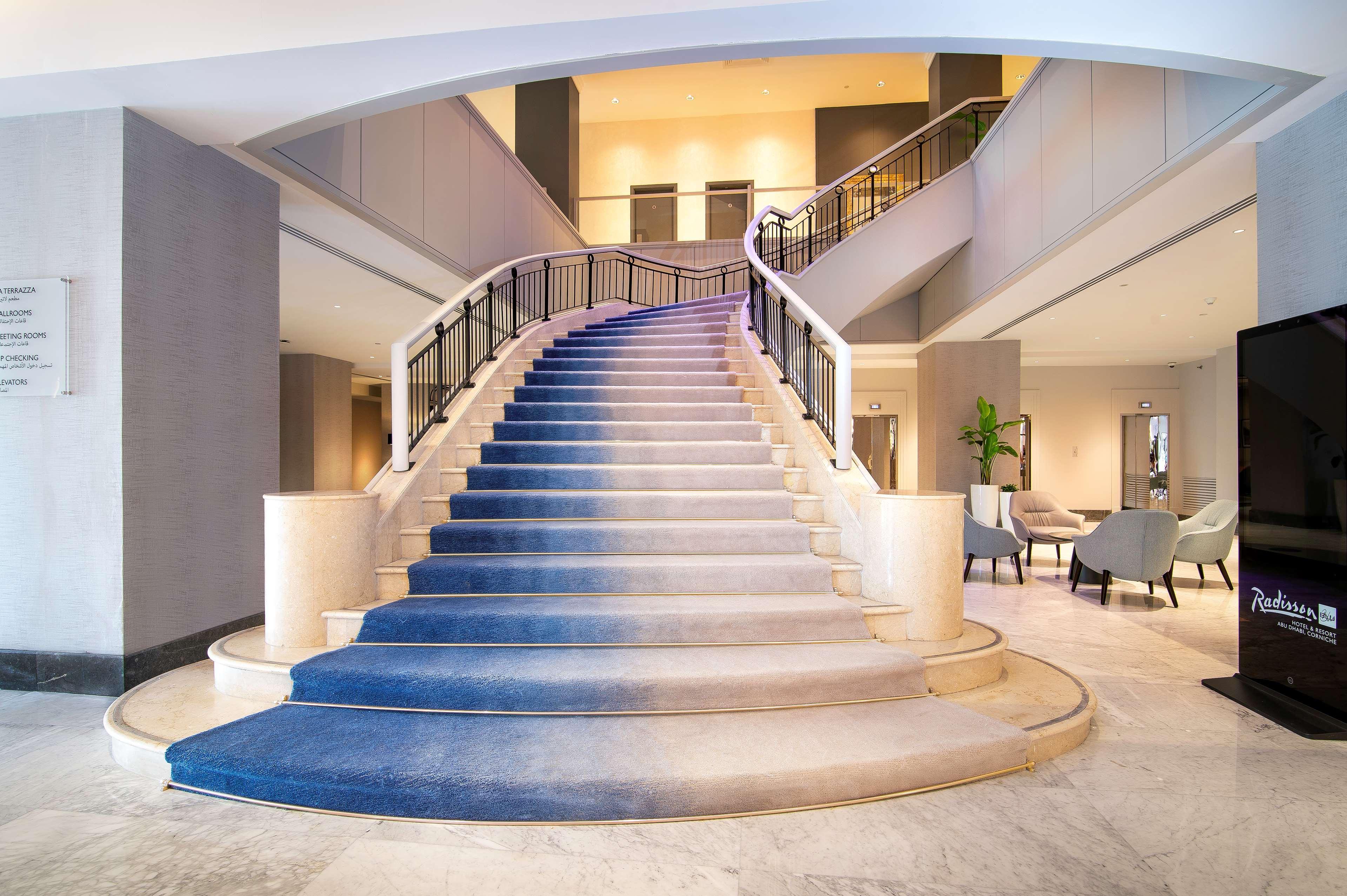 Radisson Blu Hotel & Resort, Abu Dhabi Corniche Exterior photo The photo depicts an elegant staircase inside a building, likely a hotel or a similar venue. The staircase is wide with a gentle curve, featuring a blue carpet that transitions in color from light to dark as it ascends. The walls surrounding the stai