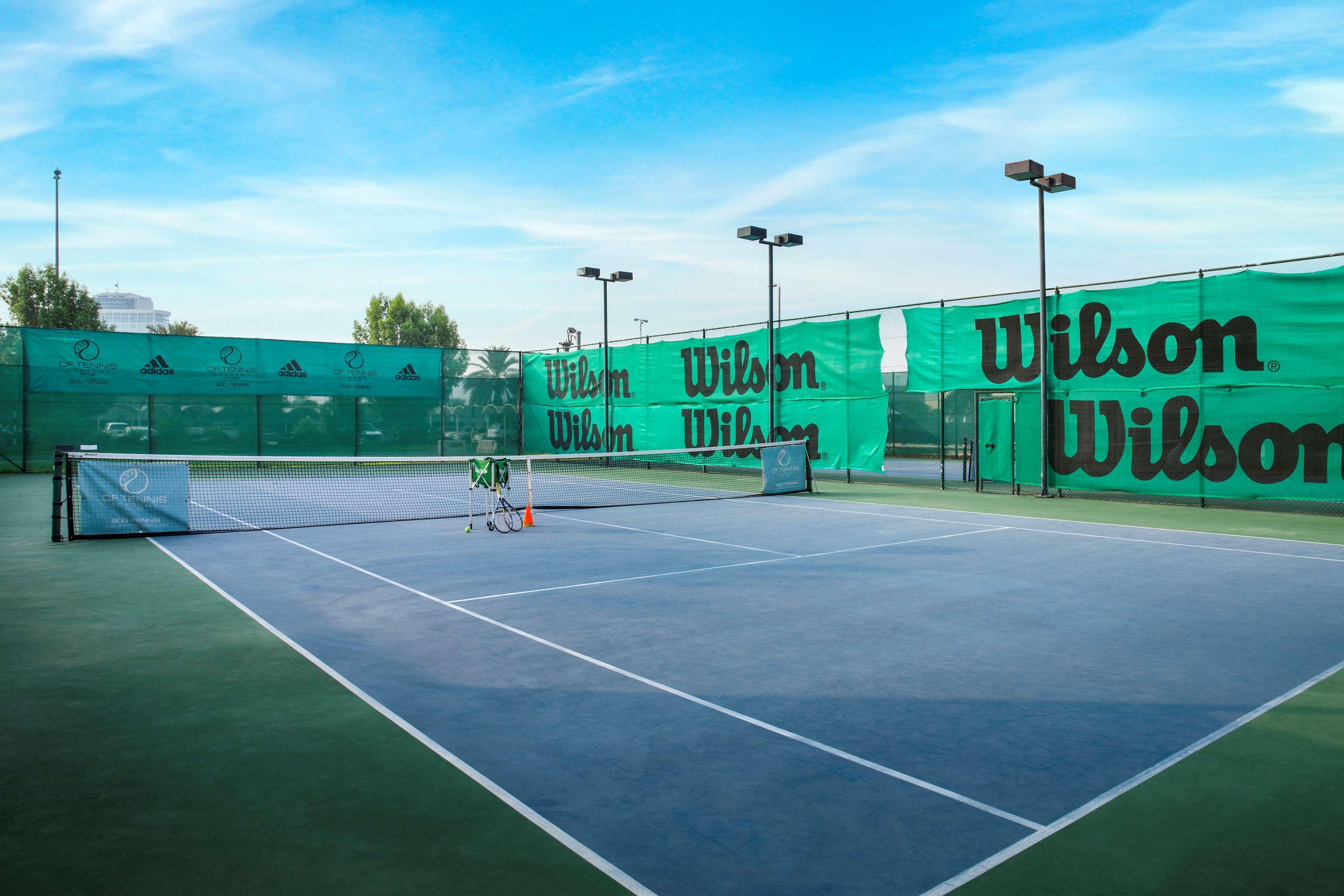 Radisson Blu Hotel & Resort, Abu Dhabi Corniche Exterior photo The photo shows a view of a tennis court surrounded by green nets and fencing featuring the brand name "Wilson." The court itself has a hard surface with marked lines for play. There are several tennis equipment items, such as a few rackets and possi