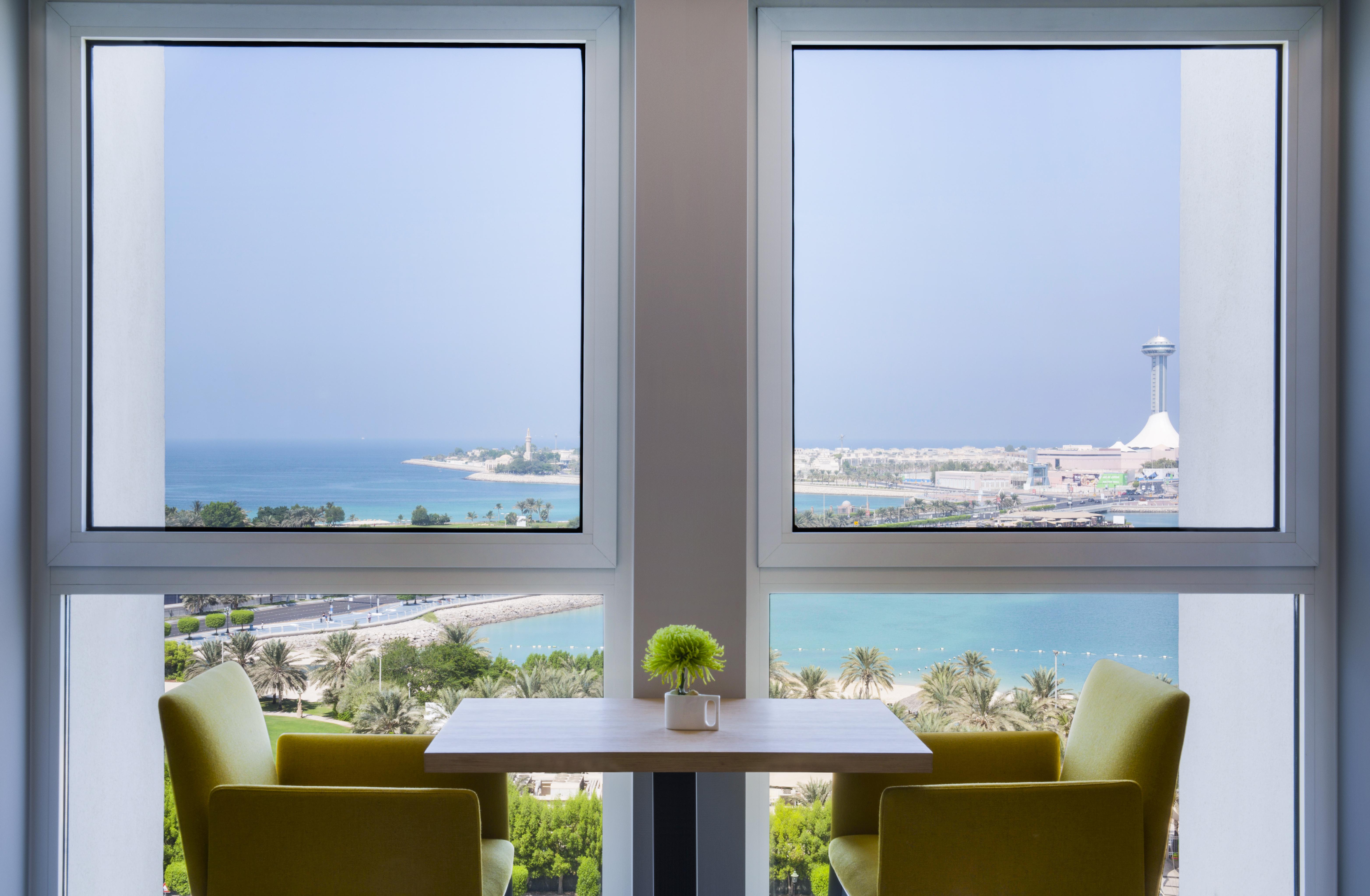 Radisson Blu Hotel & Resort, Abu Dhabi Corniche Exterior photo The photo shows a serene interior space featuring two large windows that frame a beautiful view of the ocean and coastline. The scene is bright, with sunlight filtering in, and there are two green chairs positioned around a small table. A small plant