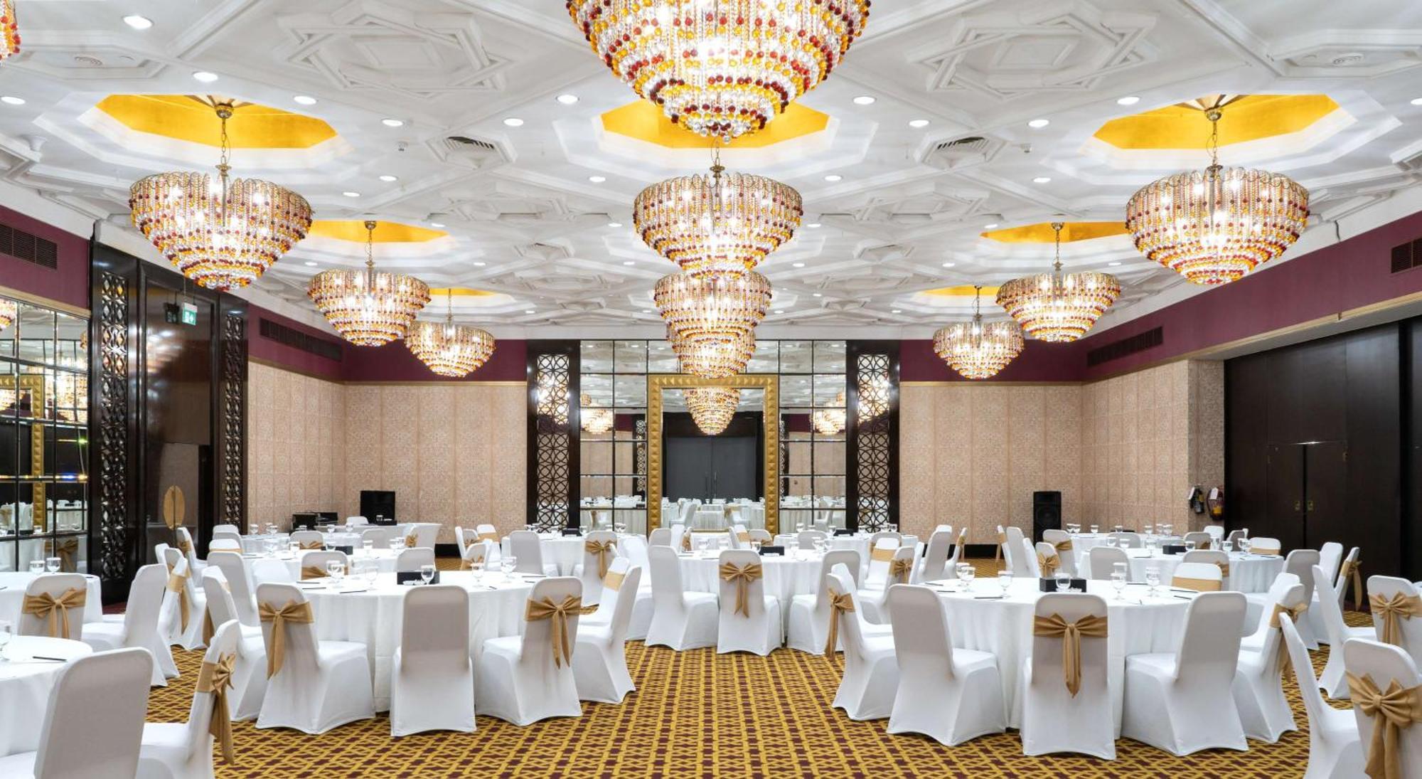 Radisson Blu Hotel & Resort, Abu Dhabi Corniche Exterior photo The photo depicts an elegant banquet hall or event space. The room is adorned with multiple large, ornate chandeliers that provide bright, ambient lighting. The tables are set with white tablecloths and chairs covered in white fabric, each decorated 